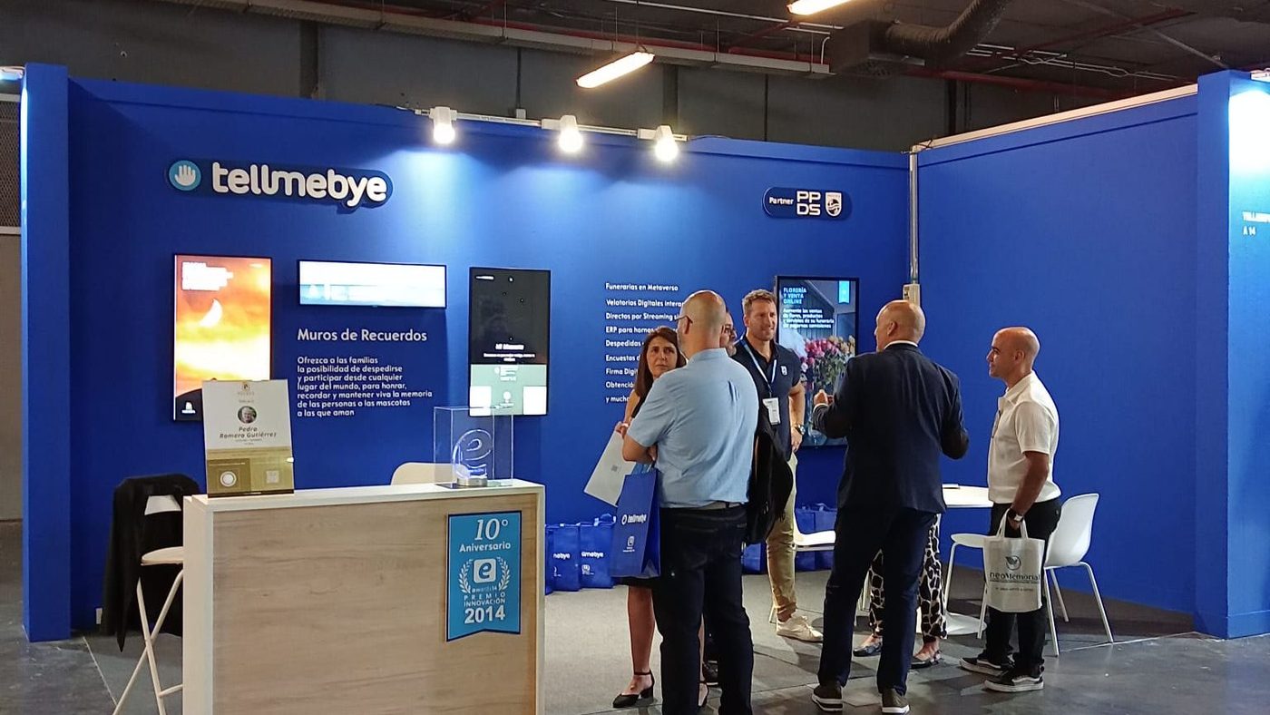 Estand de Tellmebye en Funermostra 2023, con el distintivo Partner PPDS en la pared y visitantes atendidos junto a las pantallas de Muros de Recuerdos.