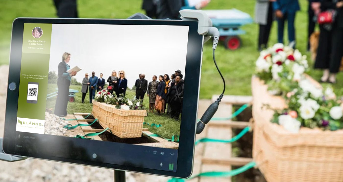 Tableta con micrófono sobre un trípode retransmitiendo en directo una ceremonia en el cementerio, con la marca de la funeraria y un código QR en pantalla.